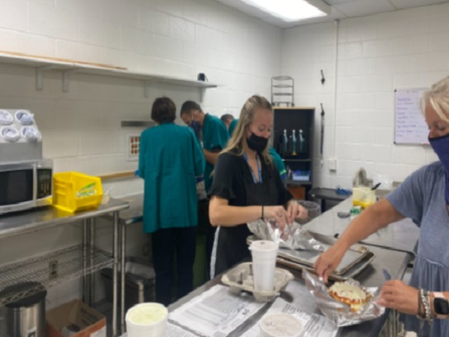 MRS. MEEKINGS ASSISTS students as they make food for this weeks Coffee Shop deliveries. The students are prepping for their usual Friday morning rush. 